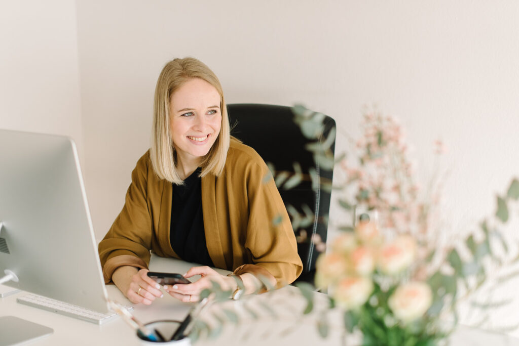 Junge, blonde Frau am Schreibtisch sitzend mit Handy in der Hand